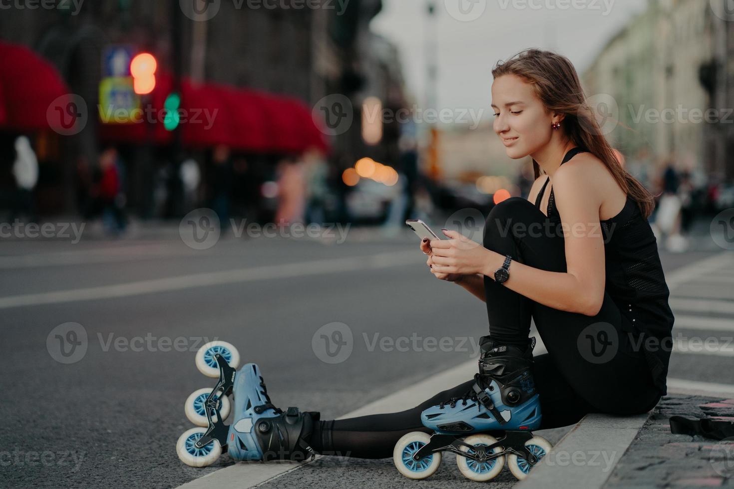 utomhusbild av sportig aktiv ung kvinna klädd i aktivt slitage bär rollerblades kontrollerar meddelandeinnehåll på smartphone poserar mot suddig stadsbakgrund på asfalt leder hälsosam livsstil foto