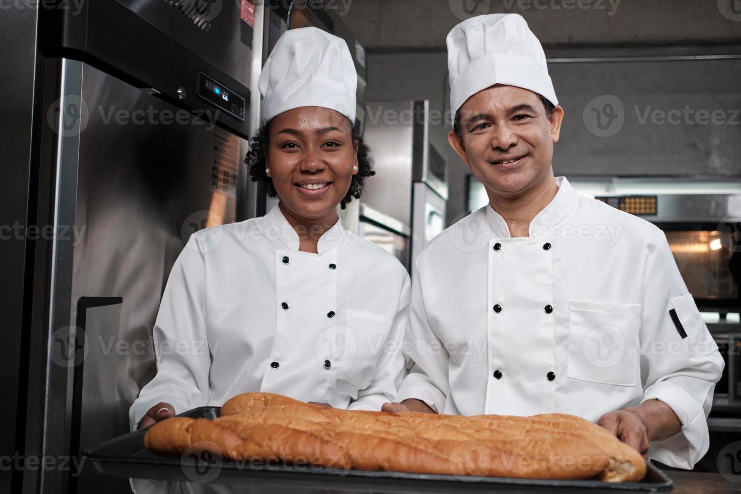 porträtt av professionella kockar i vit uniform tittar på kameran med ett glatt leende och stolt med en bricka med baguette i köket. vän och partner av bakverk och färsk daglig bageri ockupation. foto