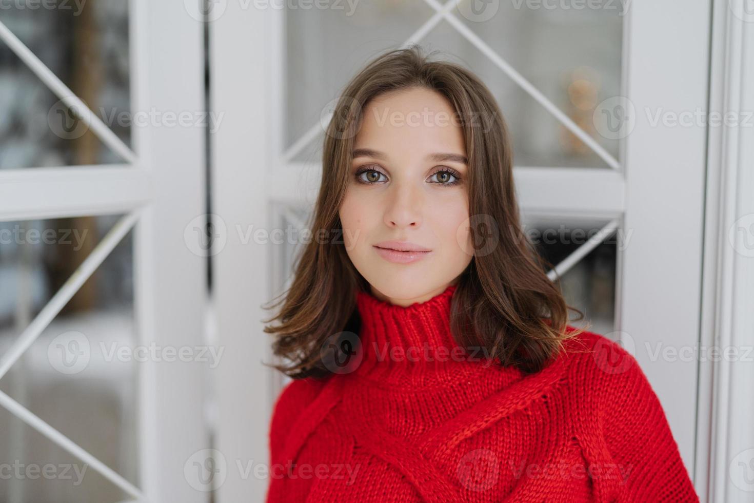 horisontell bild av lugn vacker ung europeisk kvinna bär varm röd tröja, har smink och frisk mjuk hud, poserar inomhus, tillbringar fritid hemma. människor, skönhet och livsstilskoncept foto