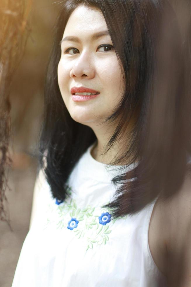 vacker asiatisk kvinna i vit klänning koppla av och ler i naturparken. Thai flicka eller kinesisk flicka njuta på semester med solljus i trädgården foto