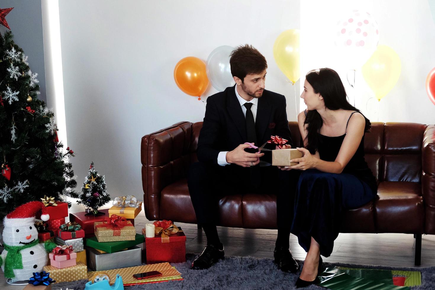 söta par älskar leende och tillbringar romantisk jultid och firar nyårsafton på brun soffdekoration med julgran, färgglad ballong och presentförpackningar i vardagsrummet hemma foto