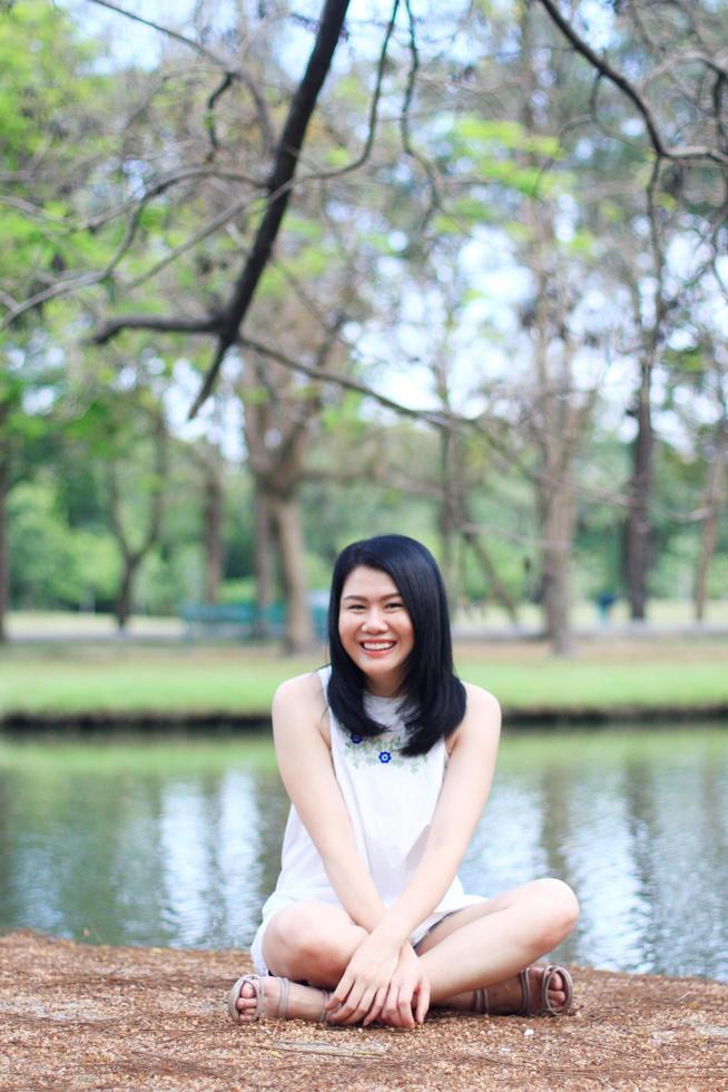 vacker asiatisk kvinna i vit klänning koppla av och ler och sitter på torrt fält nära sjön i naturparken. ung thai flicka njuta på semester med solljus i trädgården foto