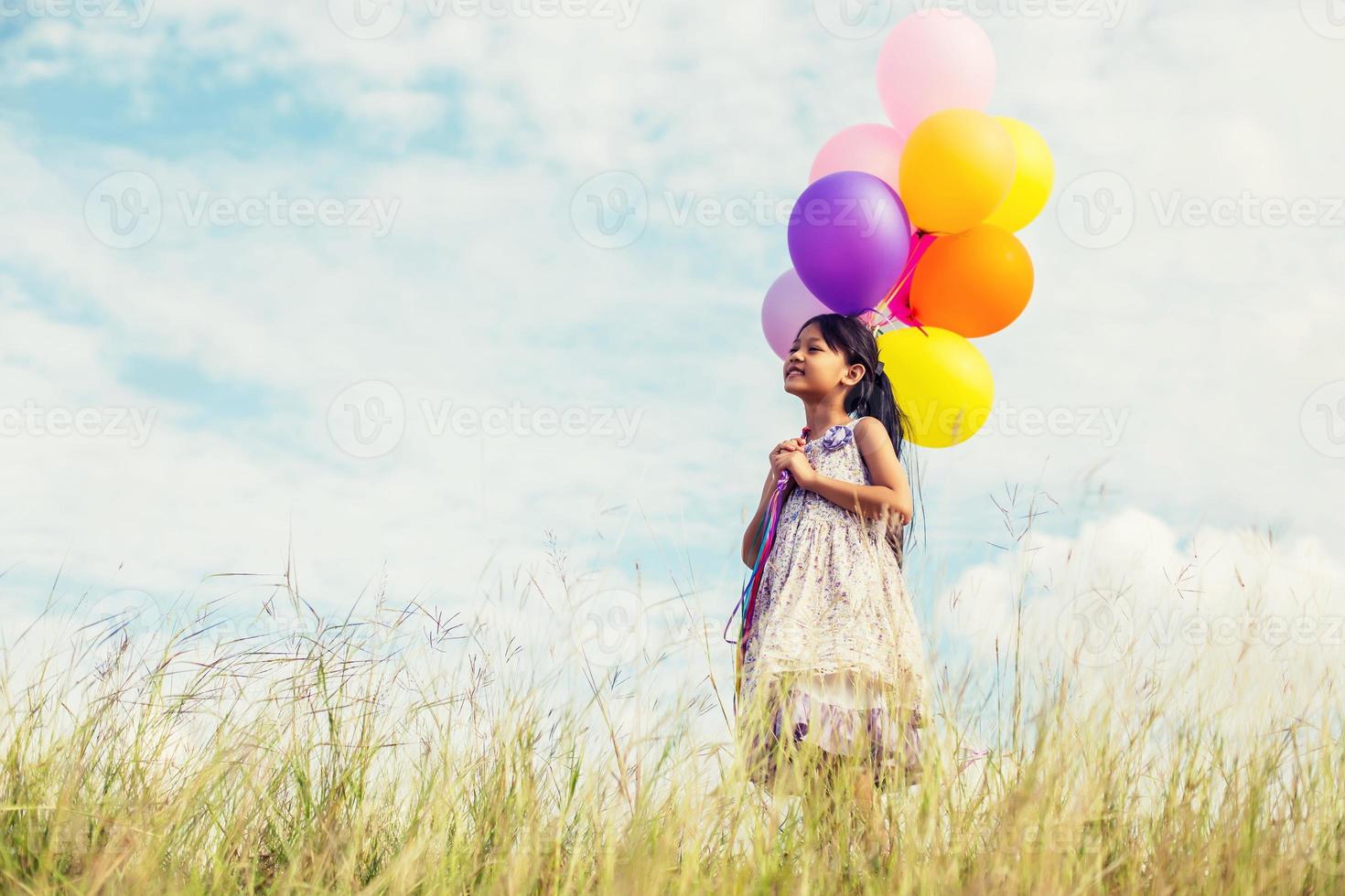 söt liten flicka håller färgglada ballonger på ängen mot blå himmel och moln, sprider händer. foto