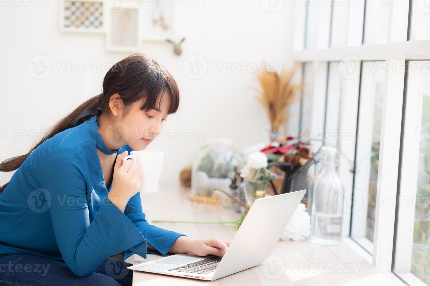 vacker ung frilansande asiatisk kvinna som ler arbetar och på bärbar dator vid skrivbordet kafé med professionell, asiatisk tjej som använder anteckningsbok och dricker kaffe, affärs- och livsstilskoncept. foto