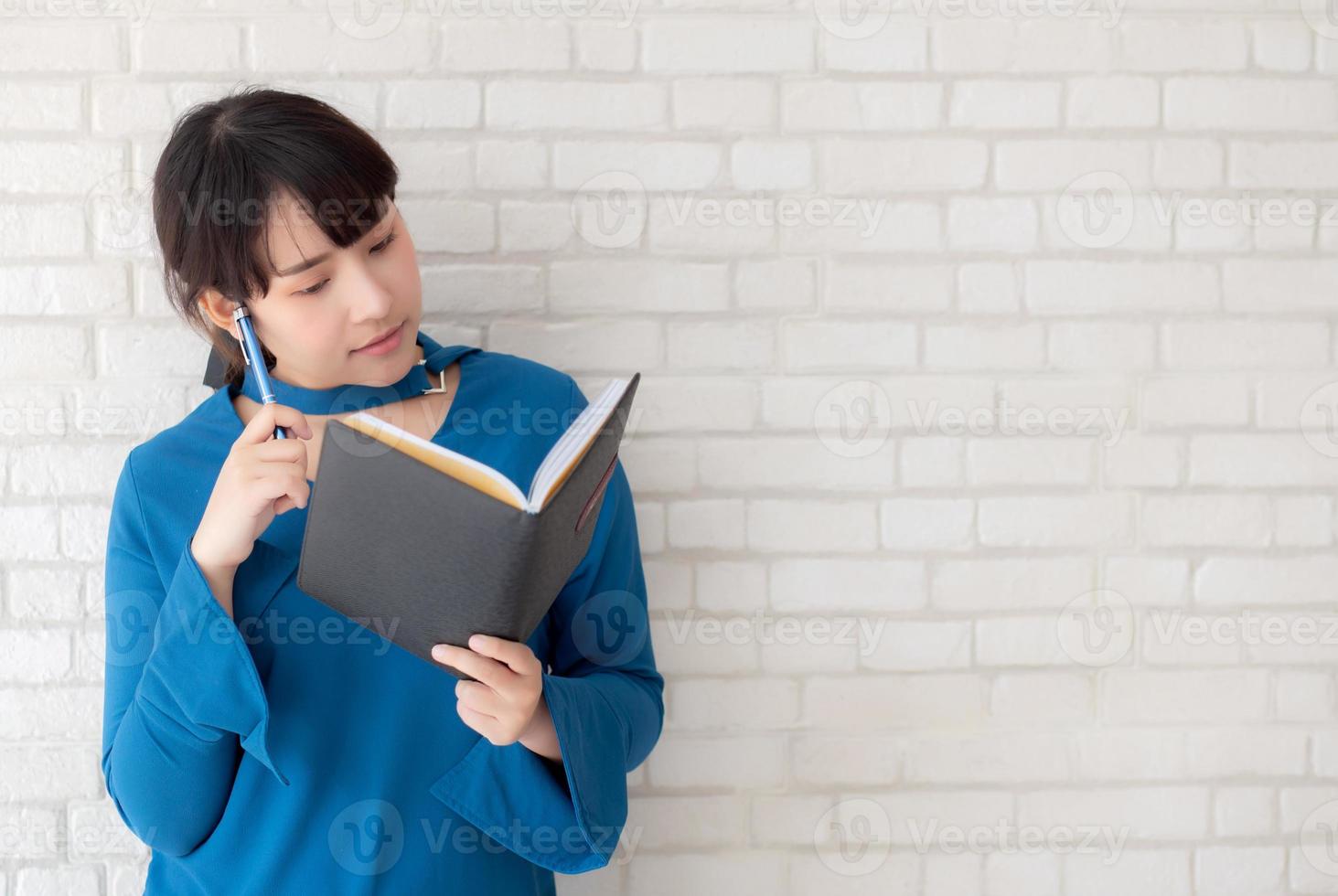 vacker asiatisk kvinna som ler står och tänker och skriver anteckningsbok på betongcement vit bakgrund hemma, flickläxa om bok, utbildning och livsstilskoncept. foto