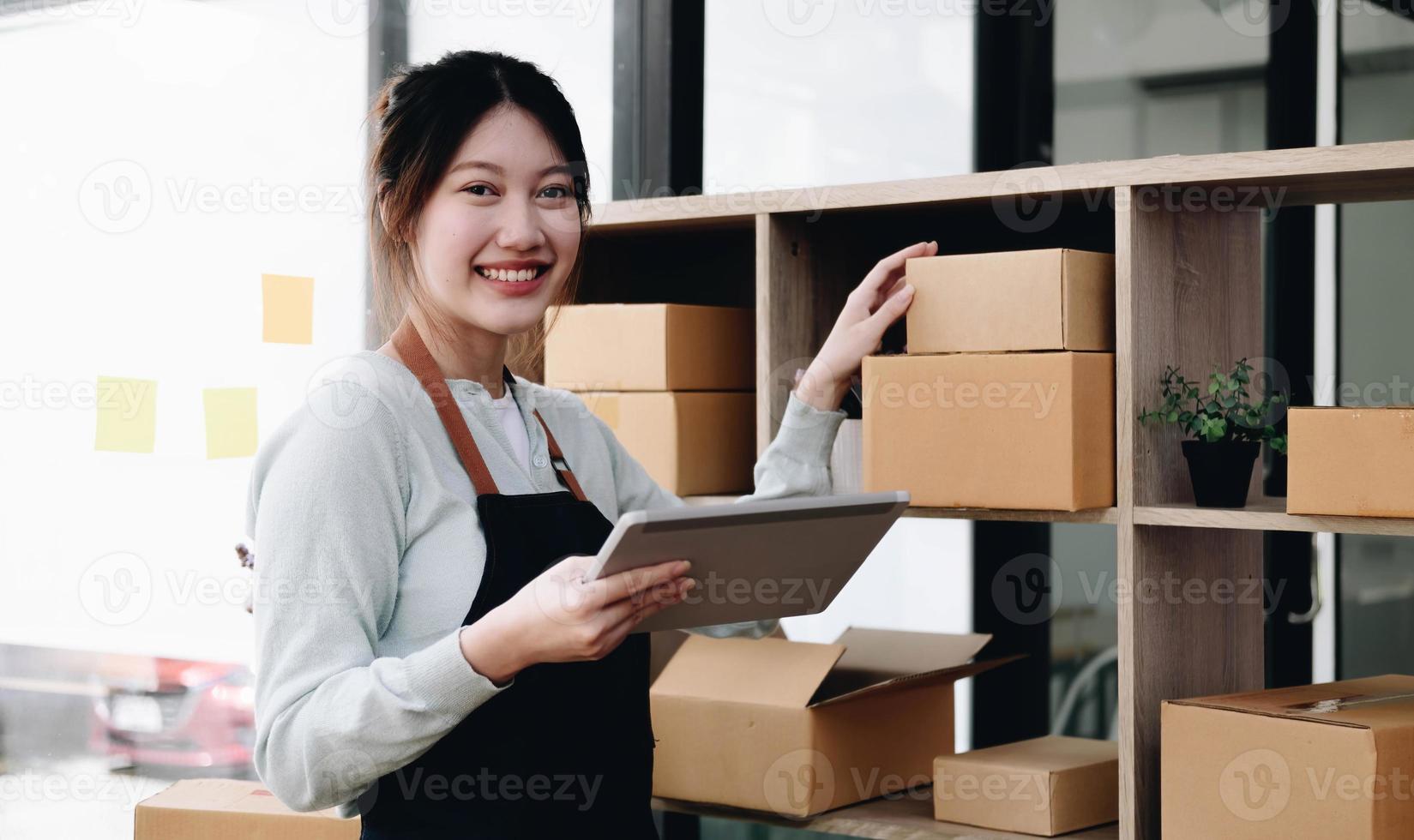 ung vacker glad asiatisk affärskvinna ägare till SM online med bärbar dator ta emot beställning från kund med paketförpackning på hennes starthemmakontor, onlineföretagssäljare och leverans foto