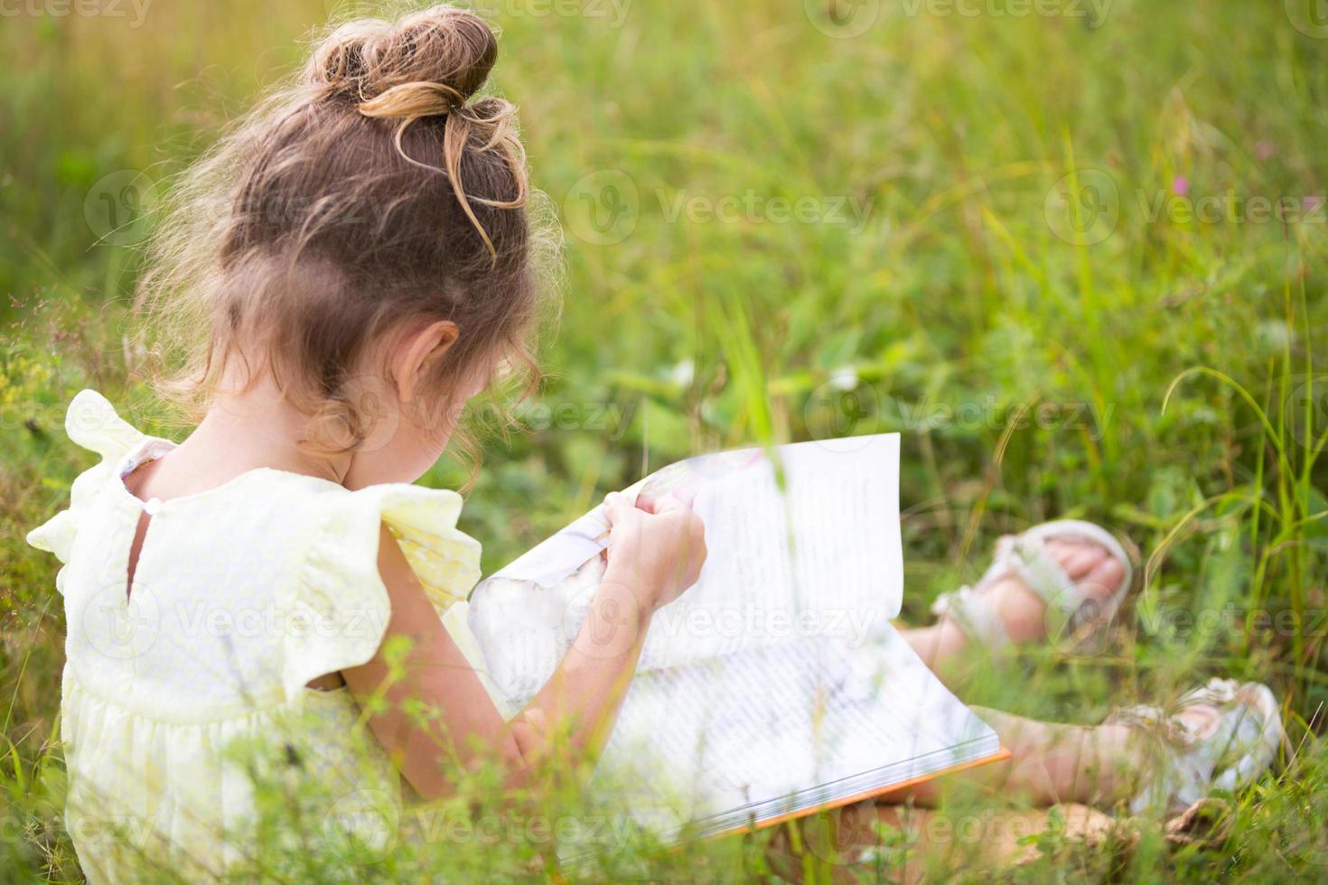 flicka i en gul klänning sitter i gräset på en filt på ett fält och läser en pappersbok. internationella barndagen. sommartid, barndom, utbildning och underhållning, stugkärna. kopieringsutrymme foto