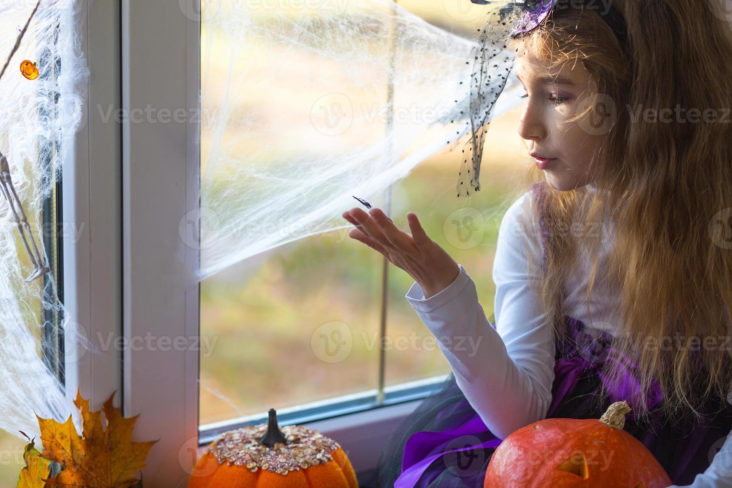en flicka i en häxdräkt på fönsterbrädan vid fönstret leker med ett spindelnät och en spindel och dekorerar huset för halloween-semestern. fruktansvärda landskap, rädsla och fasa, pumpor foto