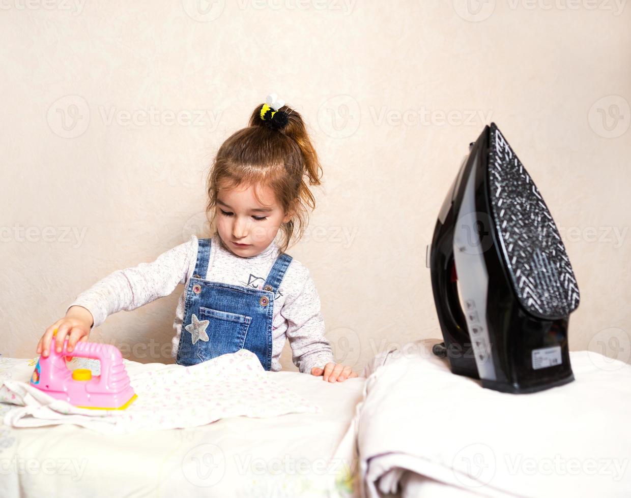 en liten flicka stryker kläder med ett leksaksjärn. mammas assistent, barnspel, hushållssysslor foto