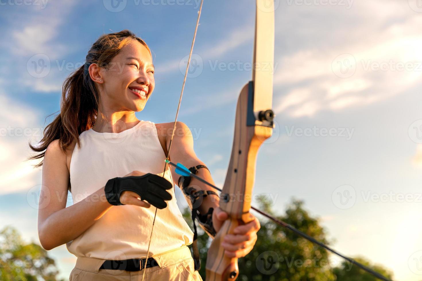 ung kvinnlig bågskytt, bågskytte, skjuta pilbåge i naturfältet till mål, framgångskoncept, på fältet för sportövning vid solnedgången foto