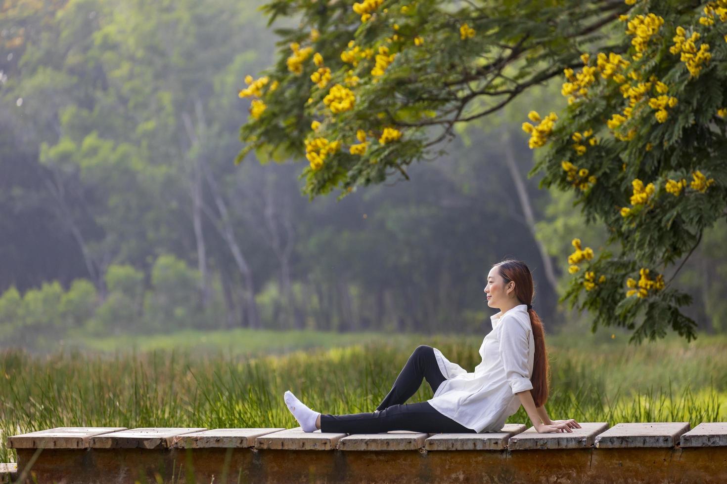 asiatisk kvinna som sitter på kanten av kajen med fridfull naturpark under sommaren med gul blomma för friluftsliv och avkoppling foto
