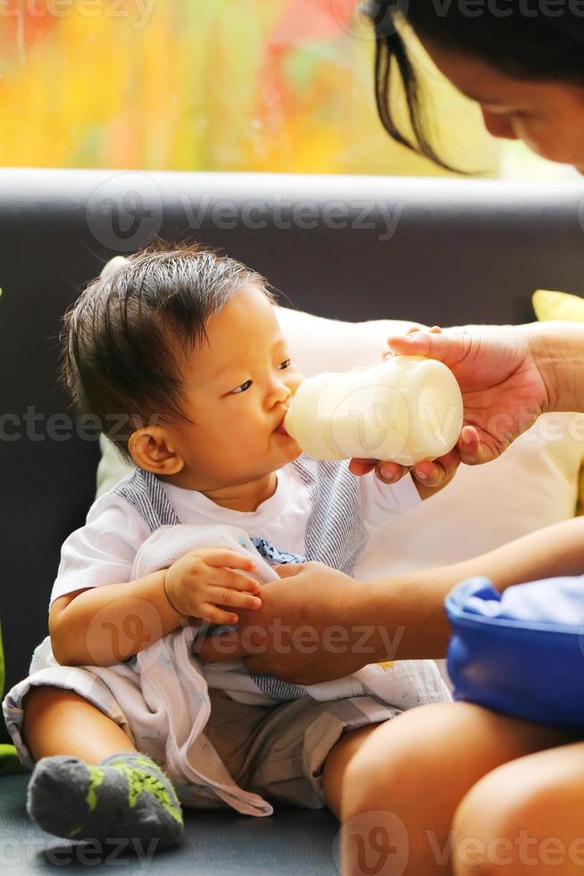 asiatisk baby sitter på soffan och dricker mjölk från nappflaska matning av mamma. foto