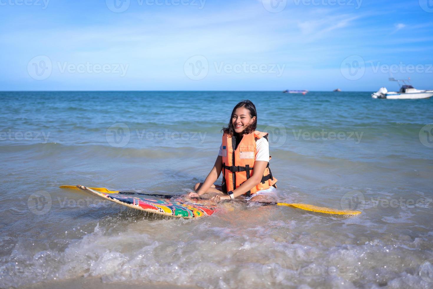 le glad asiatisk kvinna med livräddningsjacka sitta på liten paddleboard och hålla åra vid vågig havsstrand foto