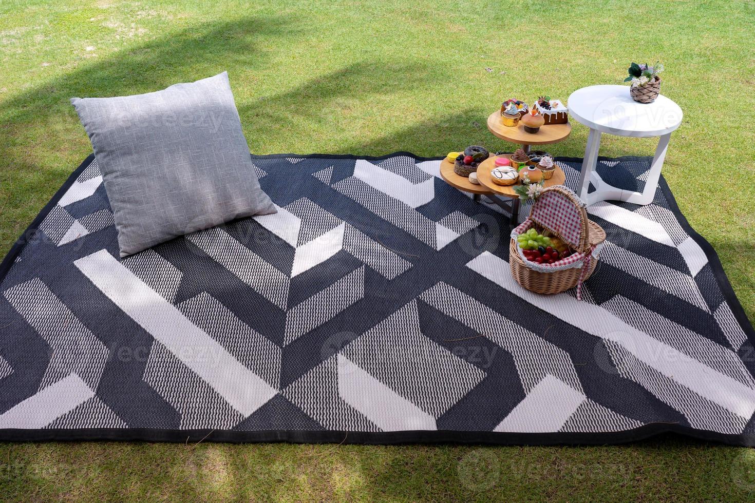 picknickset med litet bord med snacks och fruktkorg på matta med kudde i trädgården på grönt gräs foto