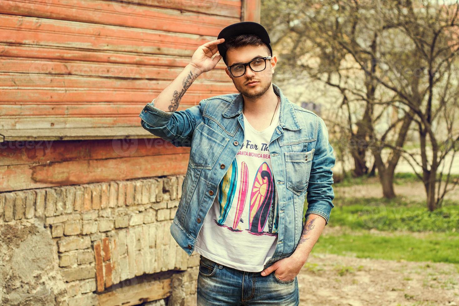 snygg stilig hipsterman i jeans med en t-shirt och solglasögon justerar sin keps och går på gatan nära ett vintage trähus foto