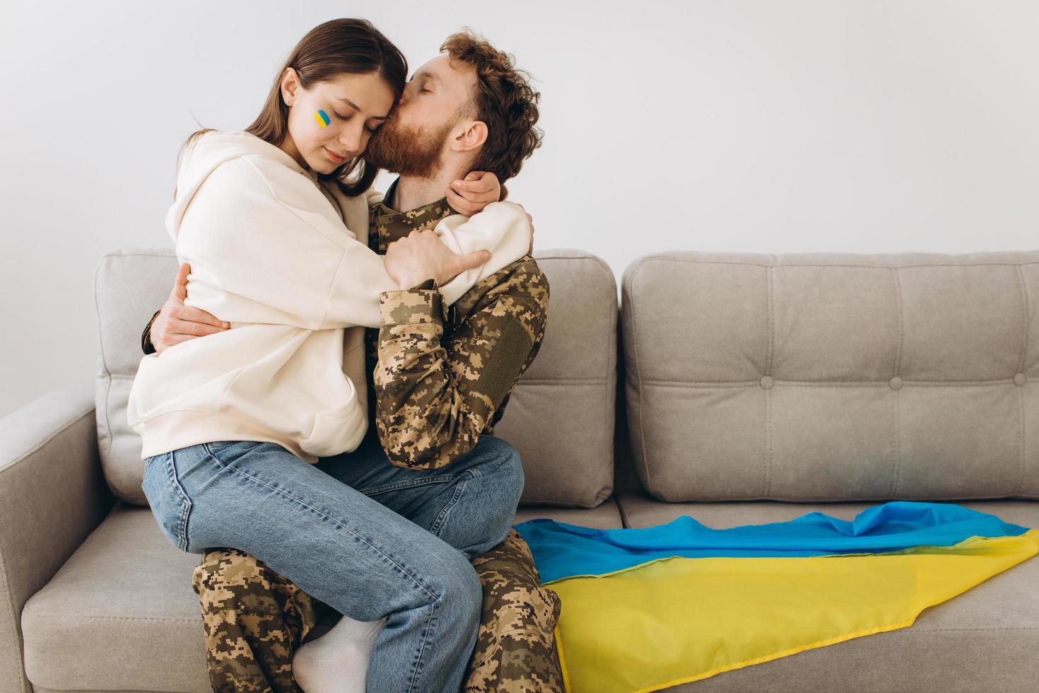 ukrainskt par, militär man i uniform kysser sin flickvän i soffan hemma på en bakgrund av gul och blå flagg foto