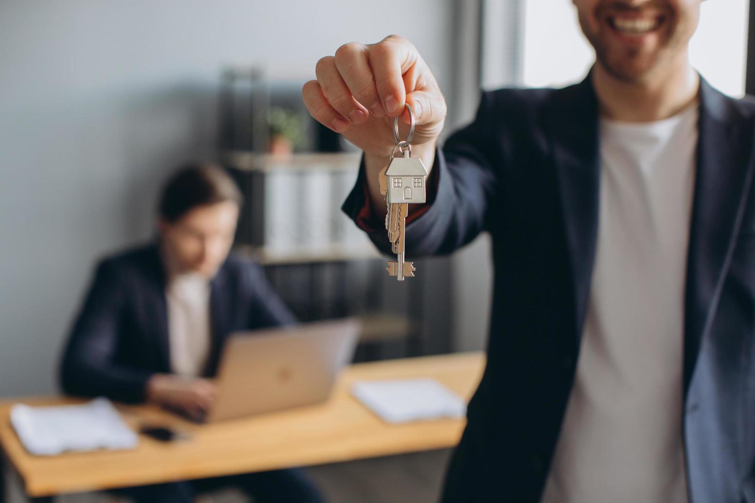leende ung modern man i kostym håller nycklarna till den köpta lägenheten i bakgrunden av mäklaren och kontoret foto