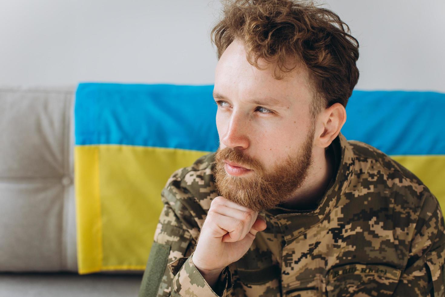 porträtt av en känslomässig ung ukrainsk patriotsoldat i militäruniform som sitter på kontoret på en soffa med en gul och blå flagga. foto