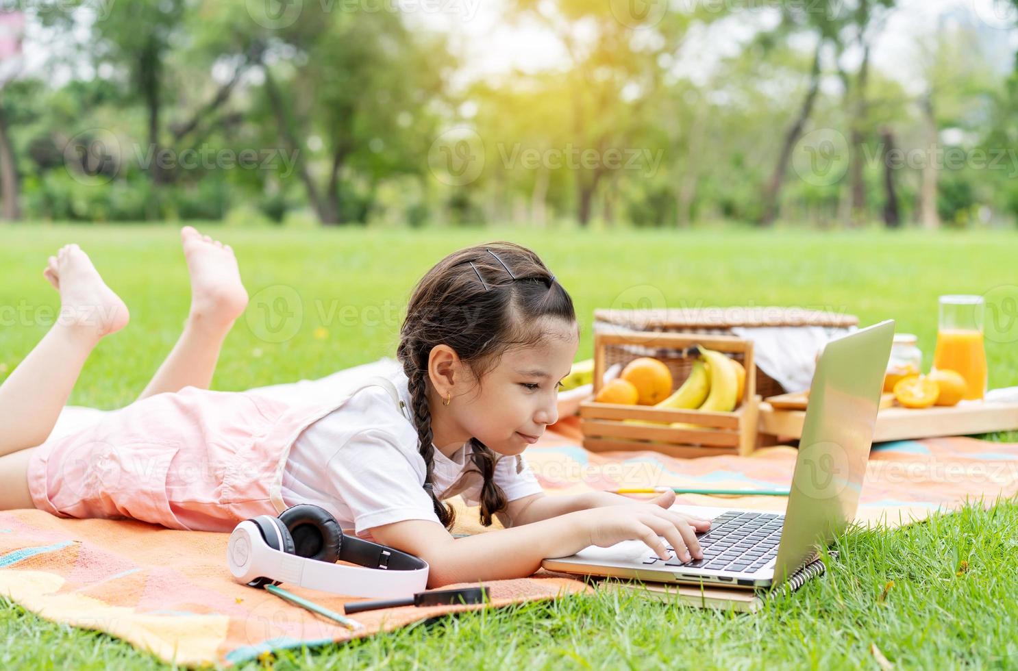 glad liten flicka barn ler, pratar, lär sig och har kul tittar på bärbar dator i sommarparken. lära sig online utbildning koncept foto