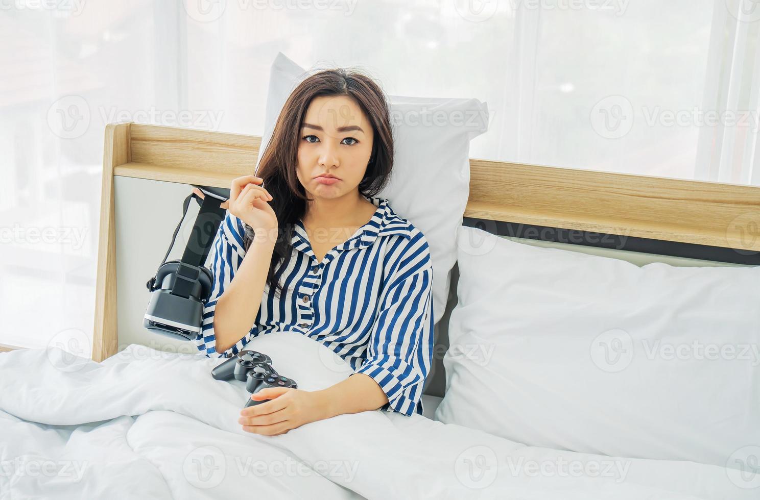 uttråkad asiatisk kvinna som håller vr-glasögon och joystick efter att ha spelat spel genom att använda vr-glasögon sittande i sängen i sovrummet, karantän på grund av coronavirus-pandemi. stanna hemma. foto