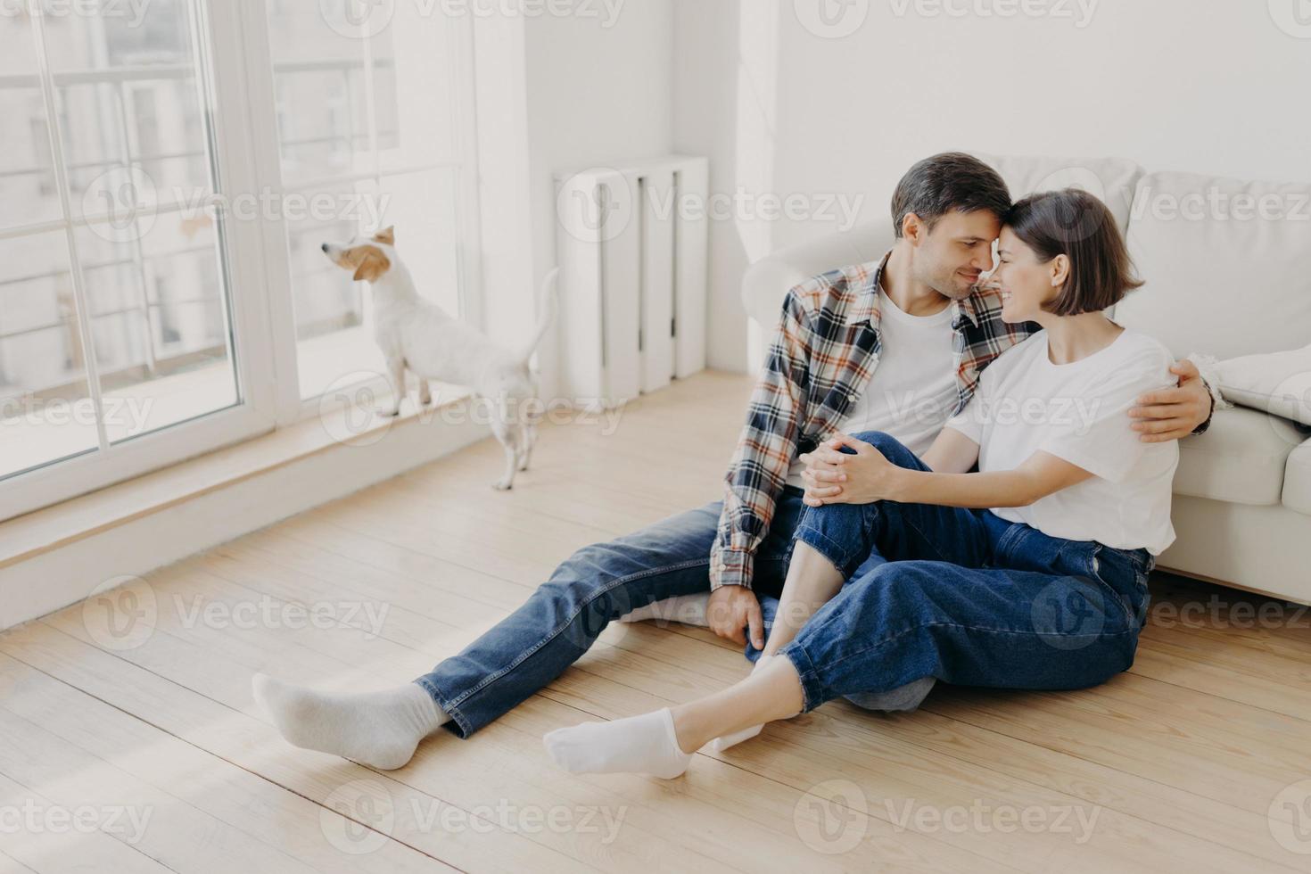 lyckligt nygift par flyttar in i ny lägenhet, gläds åt att ha fritid tillsammans, omfamnar och rör näsor, ska ha en söt puss, posera på golvet i tomt rum nära soffan, deras hund i bakgrunden foto