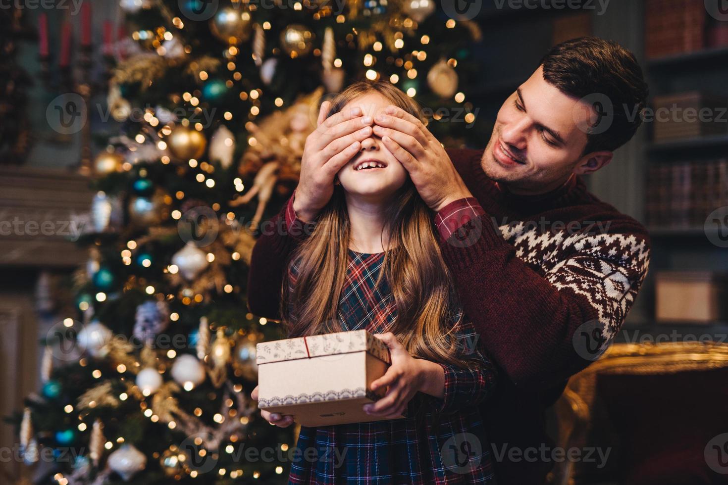 porträtt av ung far täcker hans dotters ögon som kommer att göra överraskning för henne, ger present, stå tillsammans nära julgran. glad leende flicka får present från pappa. överraskning koncept foto