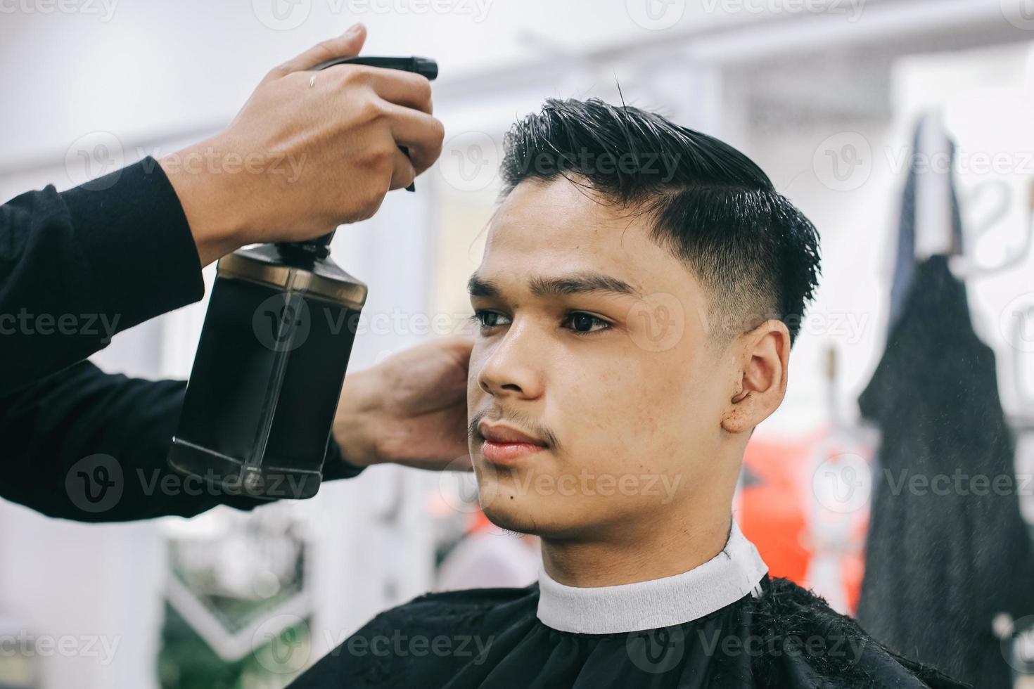 unga på frisersalongen. närbild av frisören som sprayar sin klients hår med fuktighetskräm i salongen. självvård, maskulin skönhet. barberare. foto