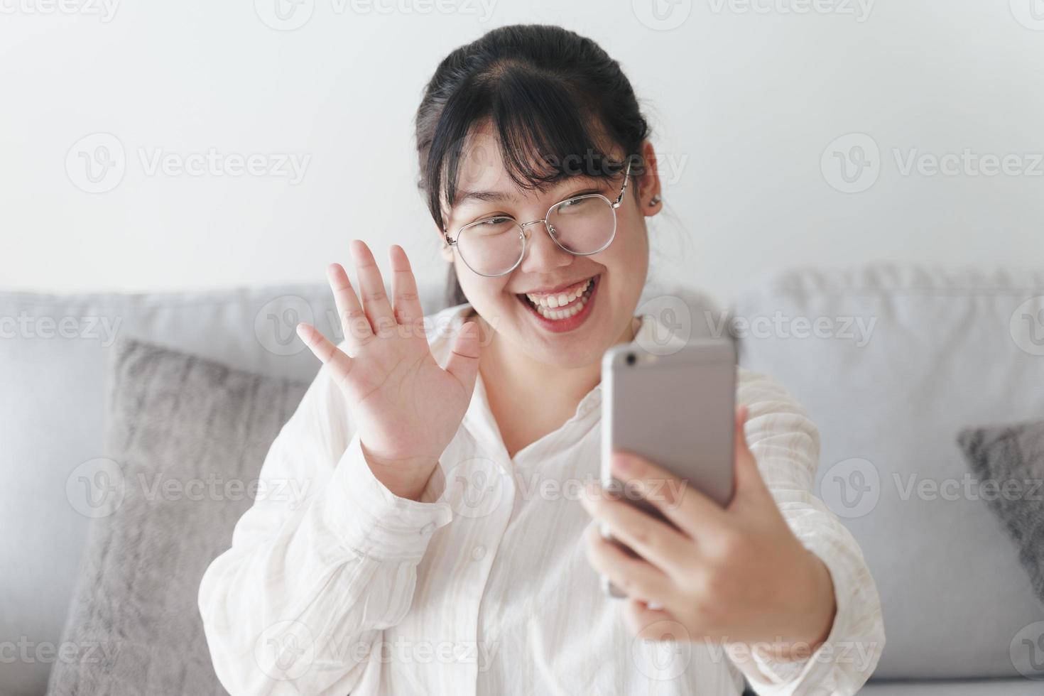 ung asiatisk kvinna som använder smartphone för online videokonferenssamtal viftande hand gör hej gest på soffan i vardagsrummet. foto