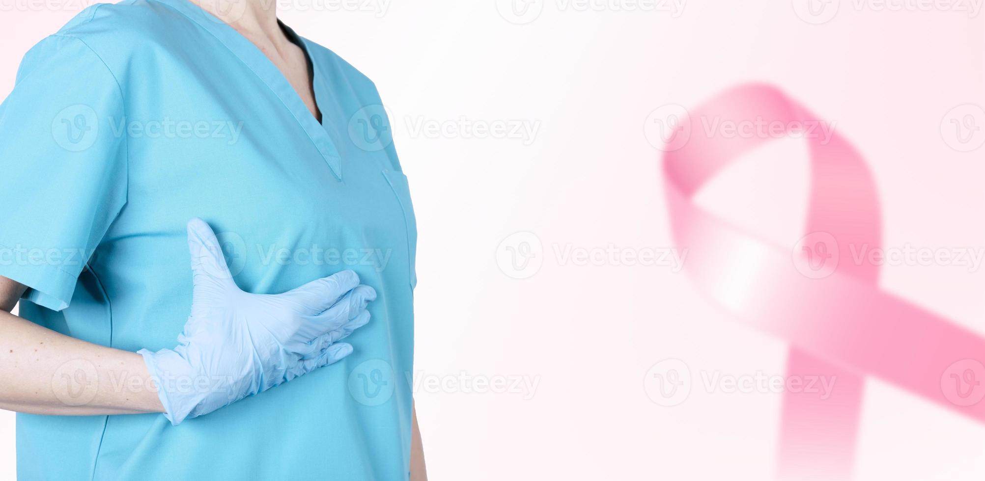 en kvinnlig läkare i medicinsk uniform stödjer hennes bröst och ett rosa band som en symbol för bröstcancermedvetenhet. medicinsk rosa bakgrund. affisch. kopieringsutrymme. foto