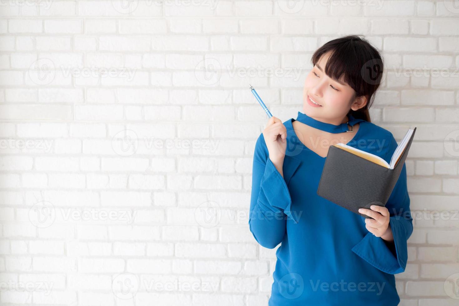 vacker asiatisk kvinna som ler står och tänker och skriver anteckningsbok på betongcement vit bakgrund hemma, flickläxa om bok, utbildning och livsstilskoncept. foto