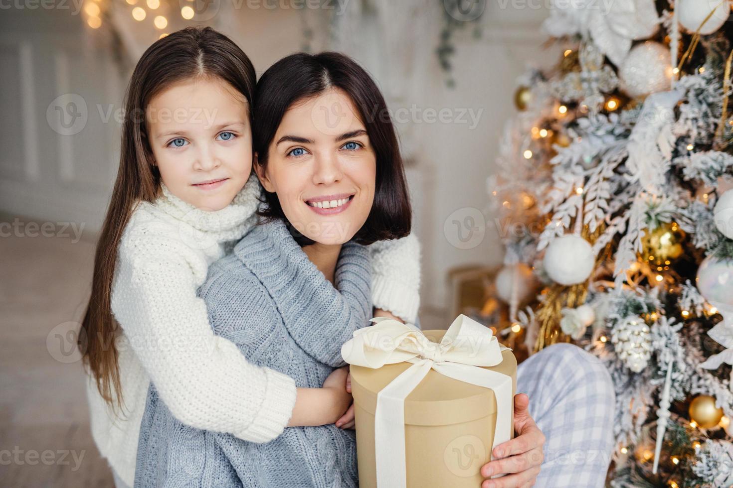 bedårande blåögd liten unge kramar med stor kärlek sin mamma som håller inslagen presentask, står nära det dekorerade nyårsträdet, glad över att fira vinterlov. människor, firande, presenterar koncept foto