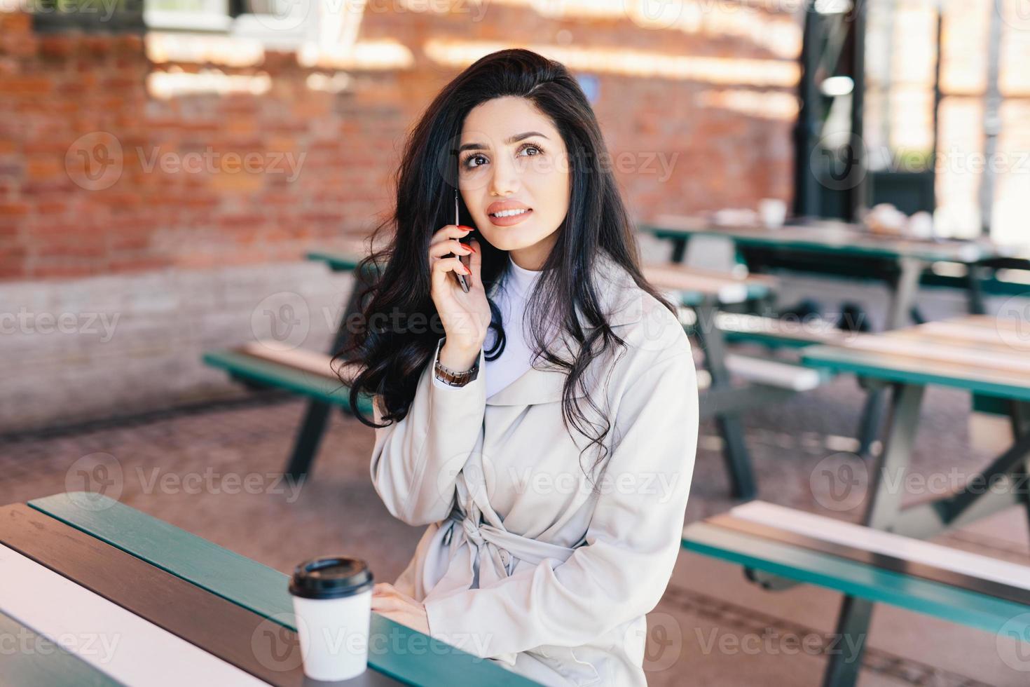 porträtt av vacker kvinna med tilltalande utseende iklädd vita formella kläder som talar över mobiltelefon och dricker takeaway-kaffe som tittar upp med ett eftertänksamt uttryck. ansiktsuttryck foto