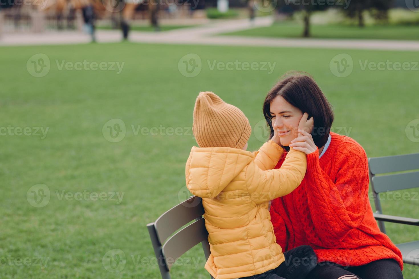 positiv ung brunettmamma bär röd tröja, har roligt tillsammans med sin lilla brorson som rör vid hennes kinder, klädd i jacka och hatt, poserar ute mot suddigt grönt gräs, sitter på en stol foto