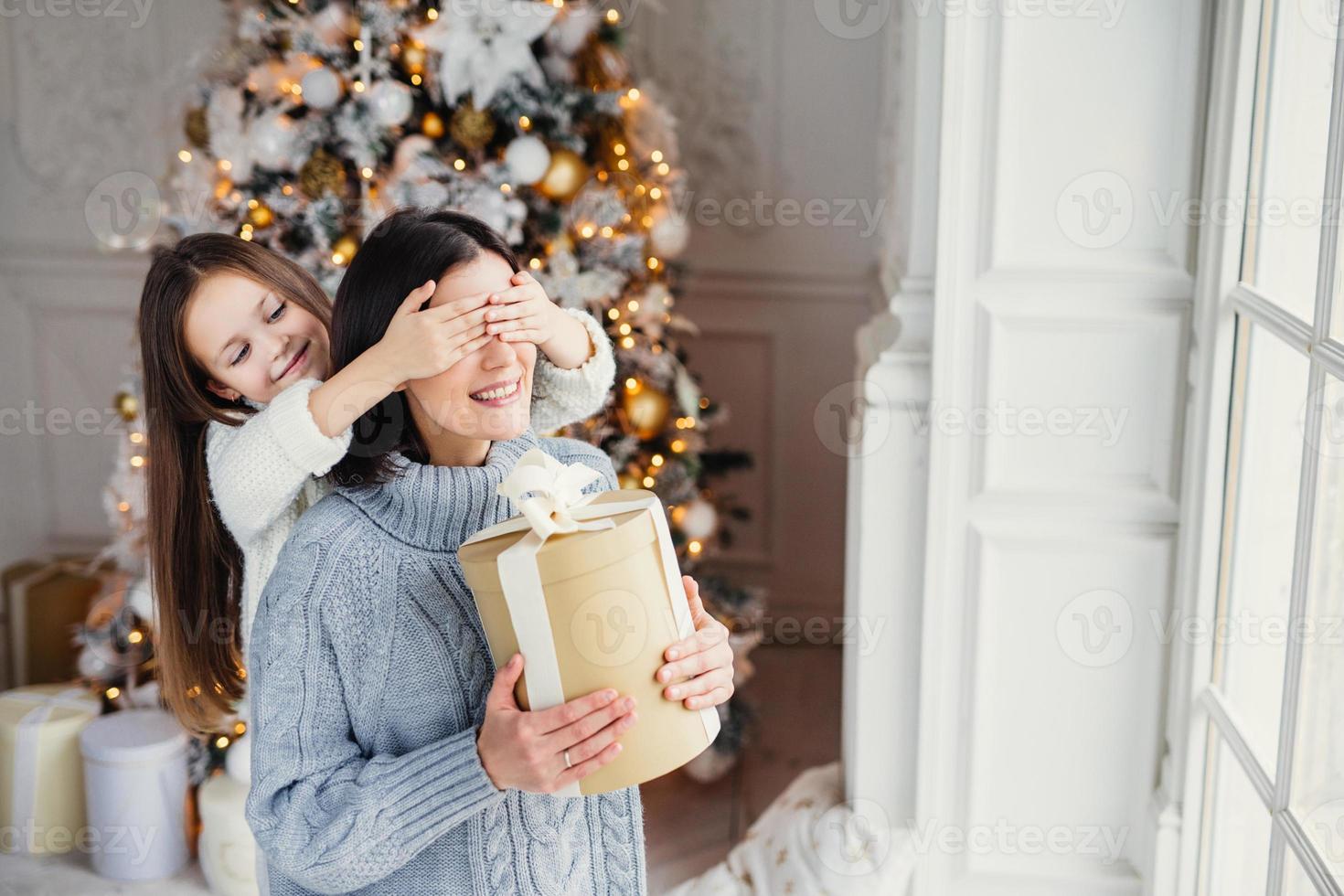 porträtt av liten flicka stänger mammas ögon, gratulerar henne med nytt år eller jul, stå nära fönstret i vardagsrummet, ha verkligt mirakel och känsla av semester. vinter, firande, årstid foto