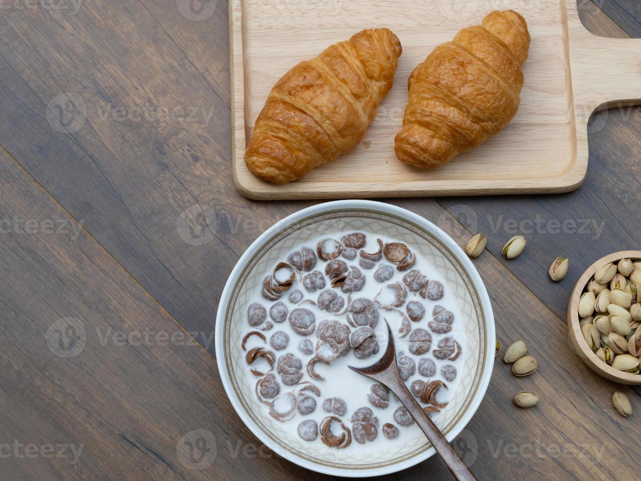 ovanifrån av frukostflingor med mjölk, croissanter och pistagenötter på träbakgrund med kopia utrymme. foto