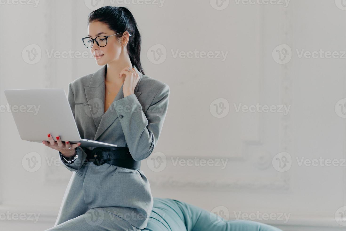 inomhusbild av seriös elegant kvinnlig administratör som jobbar hemifrån, poserar med bärbar dator, tittar på webbseminarium eller läser artikel på internet, lutar sig mot soffan, klädd i snygga affärskläder foto