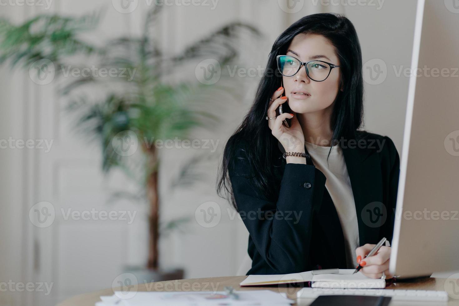 elegant ung kvinna i formell outfit svarar på telefonsamtal, skriver ner nödvändiga anteckningar i anteckningsblocket, tittar bort med tankeväckande uttryck, arbetar på en modern arbetsplats, har telefonsamtal foto