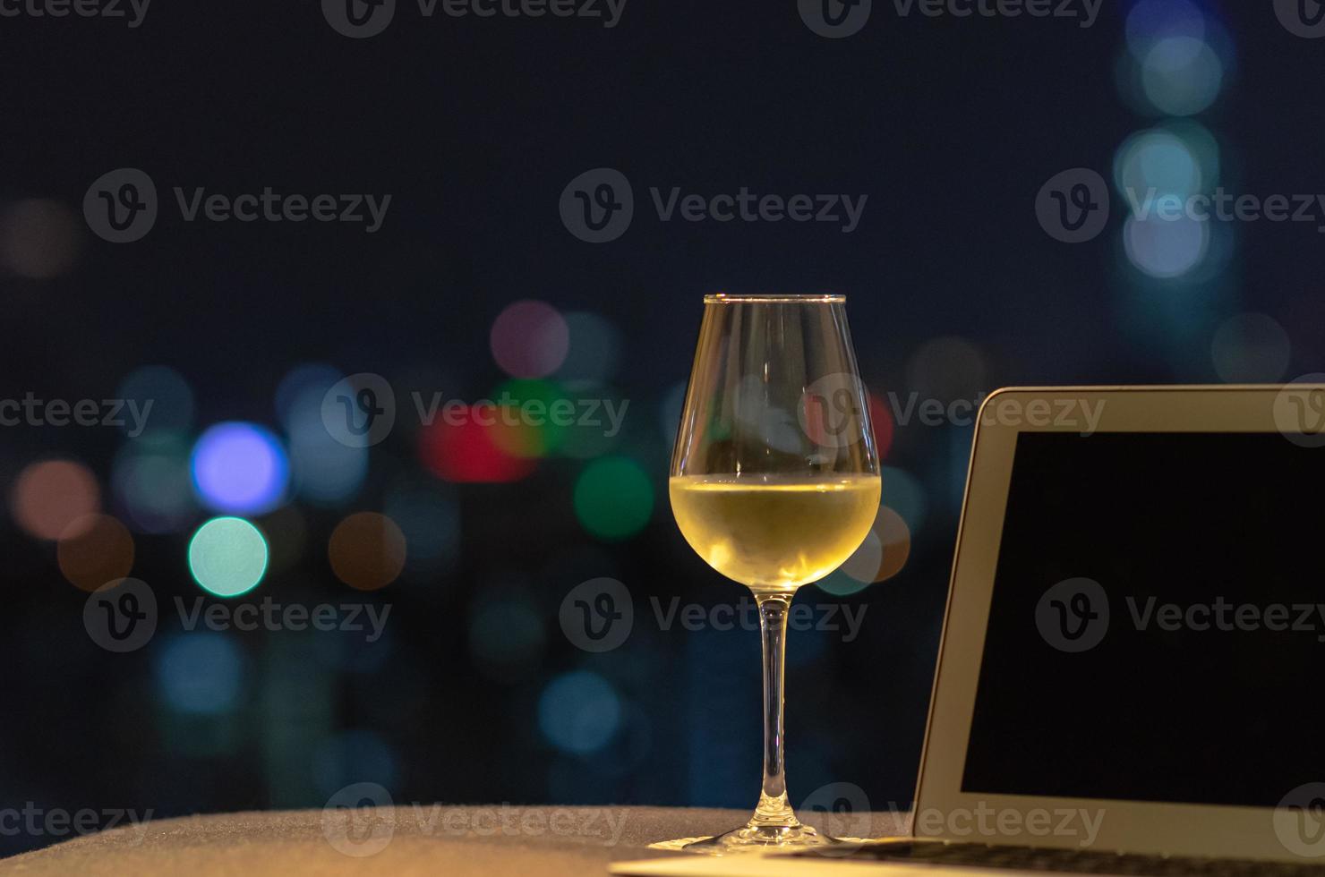 ett glas med vitt vin sätter på soffan med bärbar dator med färgglada bokeh ljus från staden. foto