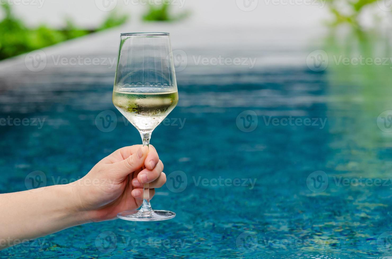 hand som håller ett glas vitt vin med poolbakgrund. semester och sommar drink koncept. foto