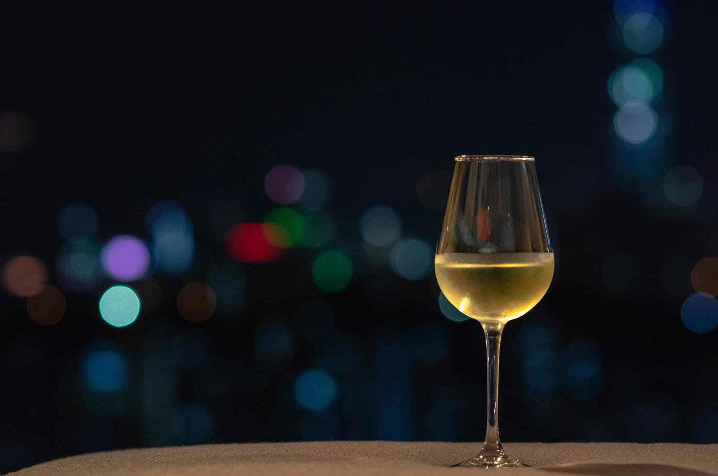 ett glas med vitt vin sätter på soffan med färgglada bokeh ljus från staden. foto