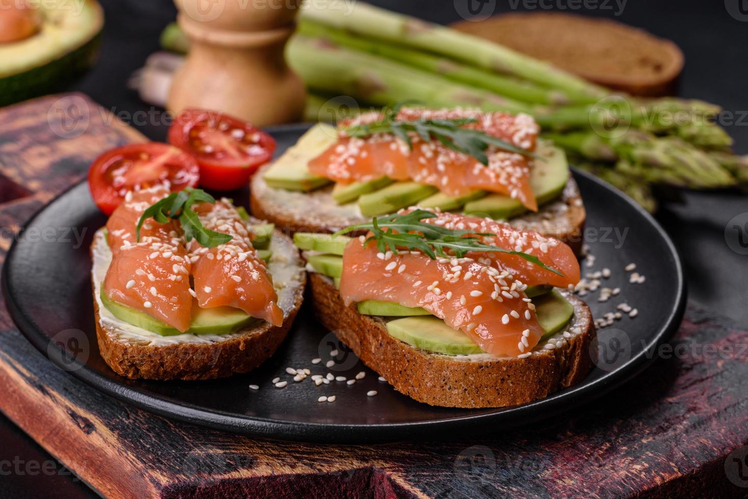 toast smörgås med smör, avokado och lax, dekorerad med ruccola och sesamfrön, på en svart stenbakgrund foto