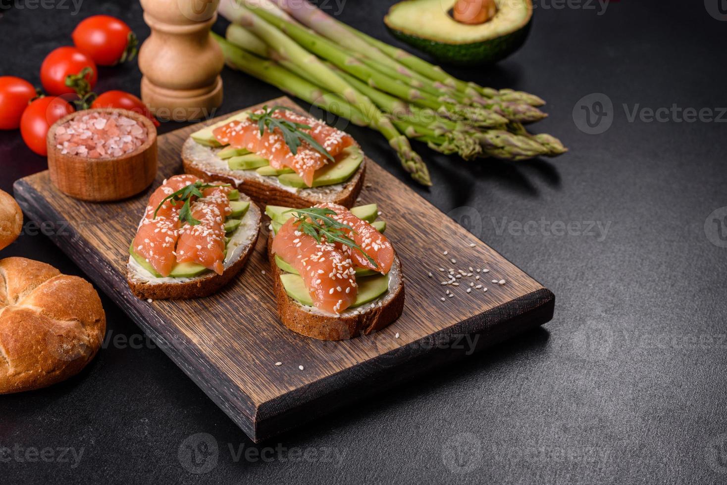 toast smörgås med smör, avokado och lax, dekorerad med ruccola och sesamfrön, på en svart stenbakgrund foto