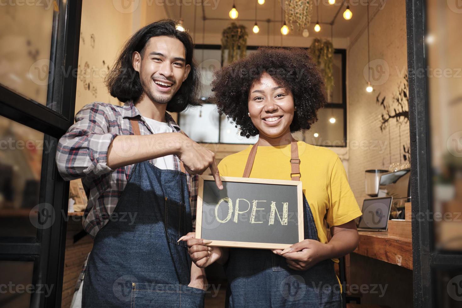 två unga startup-baristapartners med förkläden står vid casual kafédörren, bokstäver ombord och visar öppna tecken, glada och glada leenden med kaféservicejobb och nya företagare. foto