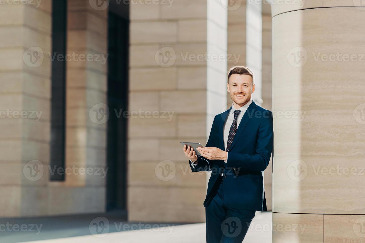 positiv framgångsrik manlig entreprenör i lyxig svart kostym, håller modern surfplatta, tittar lycklig någonstans, står i modern kontorsbyggnad, söker på internet eller sociala nätverk under arbetsuppehåll foto