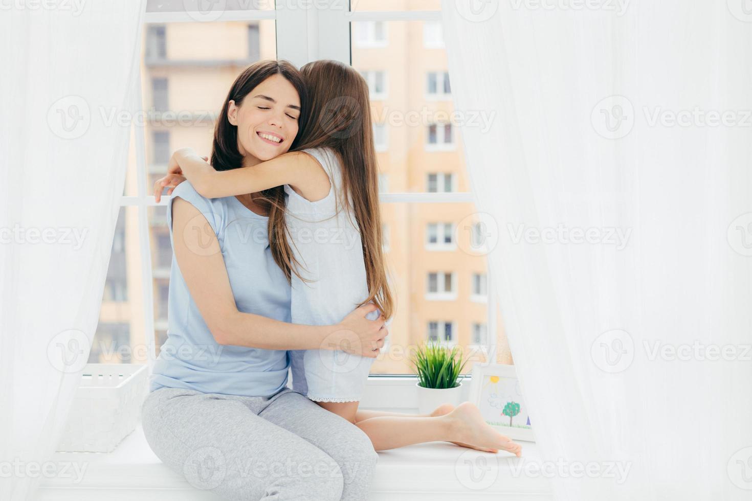 inhemsk atmosfär och föräldraskap koncept. glad mamma omfamnar sitt lilla kvinnliga barn, bär vardagskläder, poserar på fönsterbrädan, har ett vänskapligt förhållande, tillbringar helgen hemma tillsammans foto