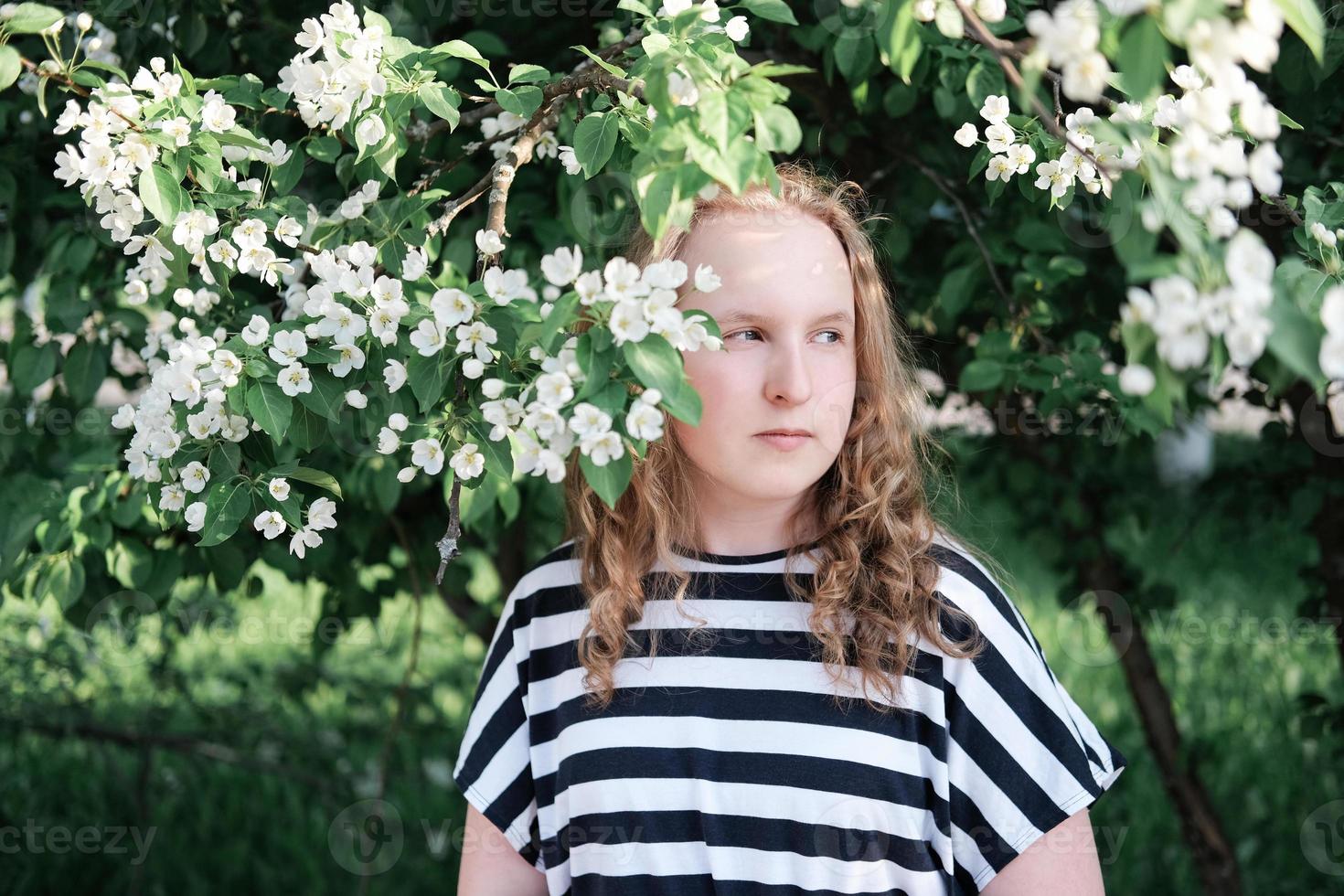 vacker tonårsflicka med lockigt hår som står nära blommande träd foto