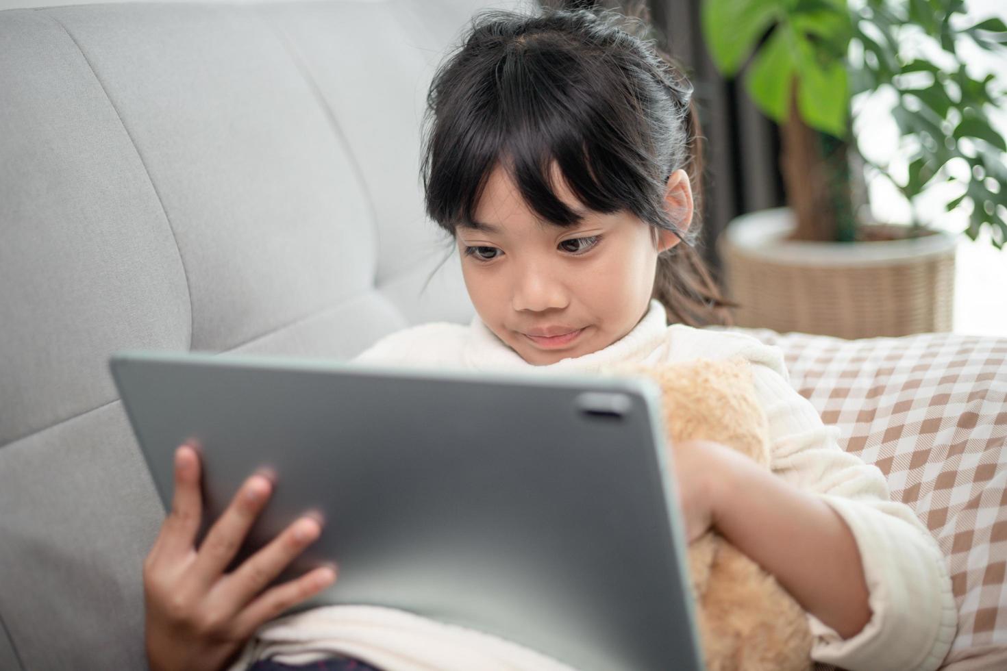 liten flicka som använder surfplatta som spelar spel på internet, barn som sitter i soffan och tittar på eller pratar med en vän online, barn kopplar av i vardagsrummet på morgonen, barn med nytt teknikkoncept foto