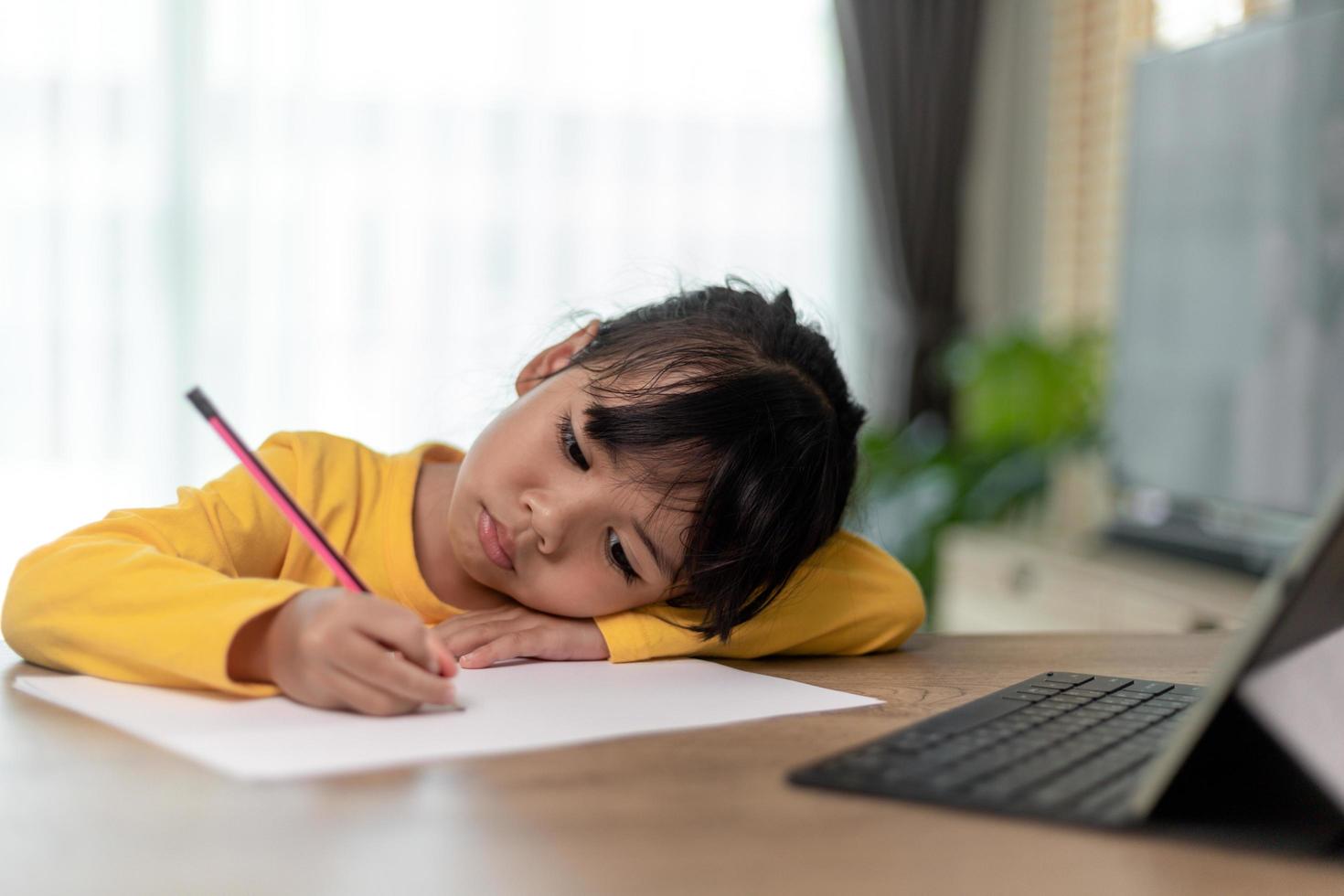 liten asiatisk tjej som sitter ensam och tittar ut med ett uttråkat ansikte, förskolebarn som lägger huvudet på bordet med ledsen uttråkad med läxor, bortskämt barn foto