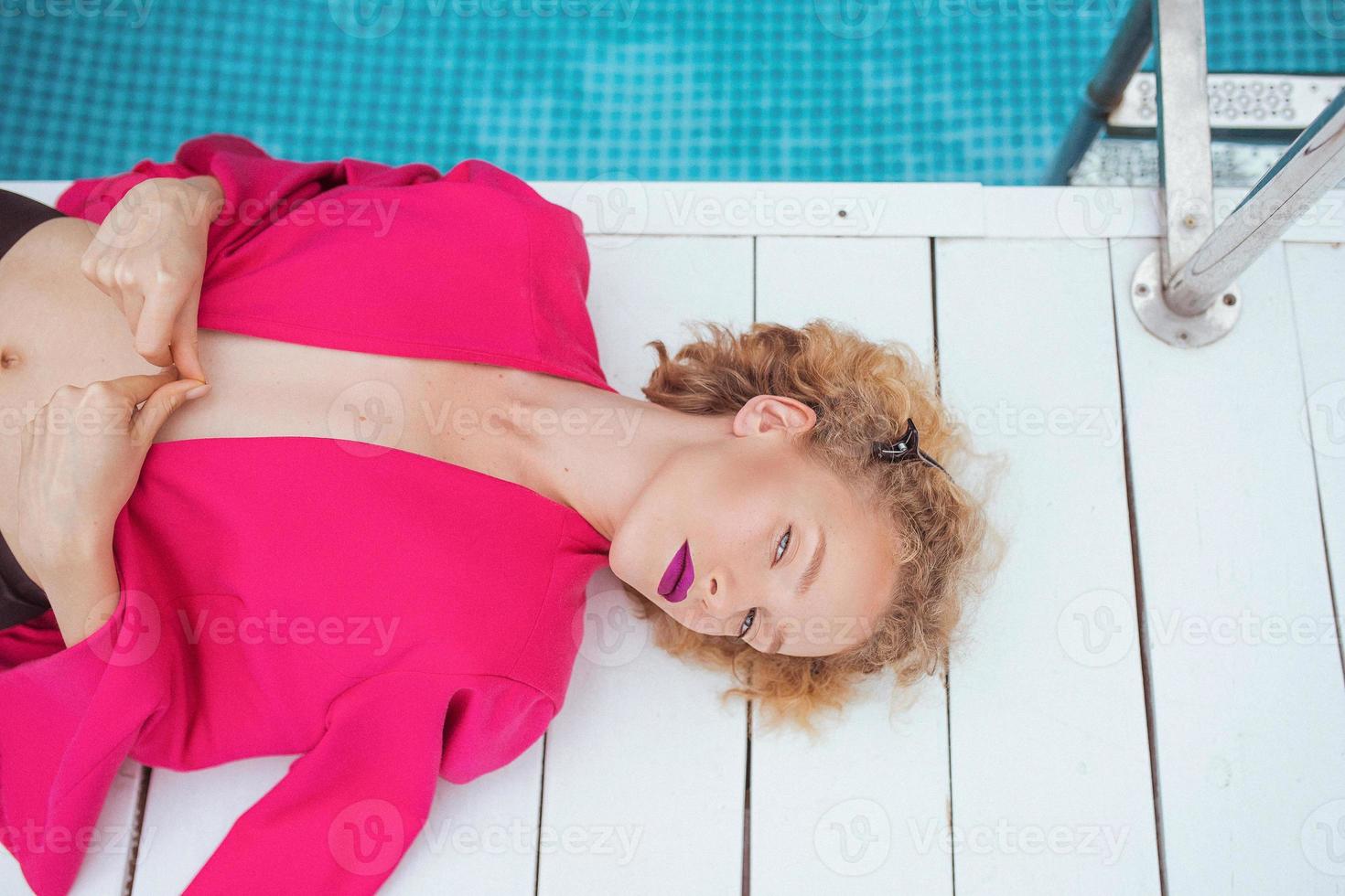 porträtt av underbart snygg rödhårig lockig ung kvinna som ligger i fuchsiajacka vid poolen. mode, stil, fritid, rekreation, resor, smink koncept. filmkorn foto