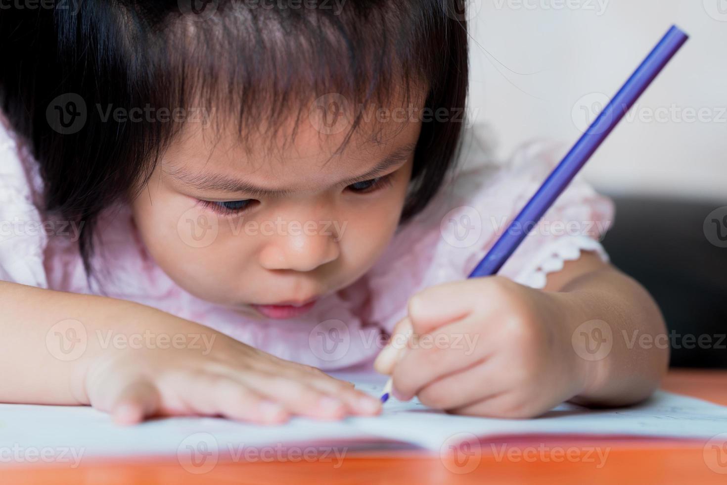närbild söt asiatisk barn flicka färgläggning i hennes läxbok. barn är villiga att lära sig på dagis. barn som håller blå penna. barn hukade nära boken för att titta närmare. foto